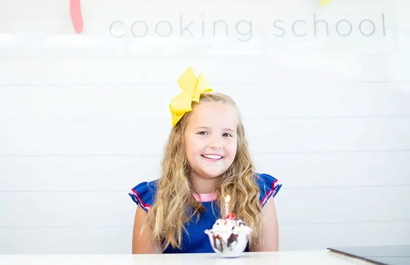 Smiling girl with yellow bow in a cooking school, holding a dessert with a candle.