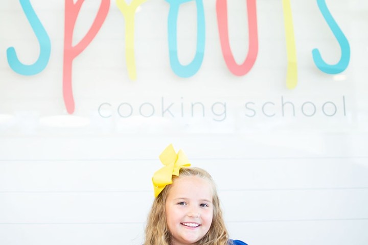 Girl with yellow bow and cupcake smiles in front of 'Sprouts Cooking School' sign.