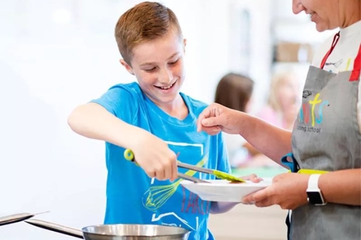 Boy in a blue shirt learning to cook with an adult, using a spatula over a stove.
