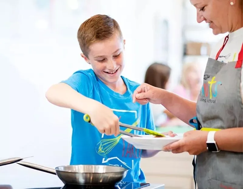 Boy in blue shirt cooking with a woman in apron, holding a plate.
