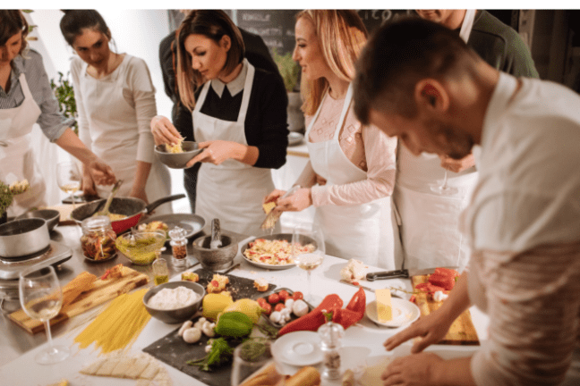Group cooking class with people preparing dishes and pasta at a kitchen counter.