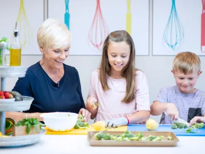 Older woman and two kids slicing vegetables together on colorful chopping boards.