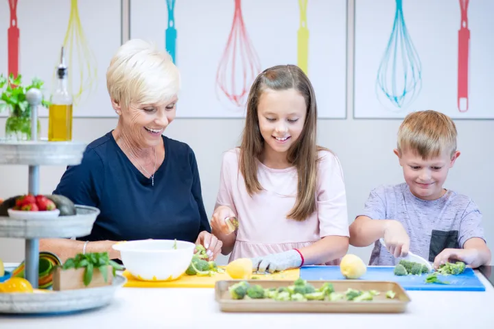 Older woman and two kids slicing vegetables together on colorful chopping boards.