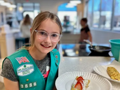 Smiling girl in glasses and green vest with rolled crepe on a plate, topped with cream and strawberries.