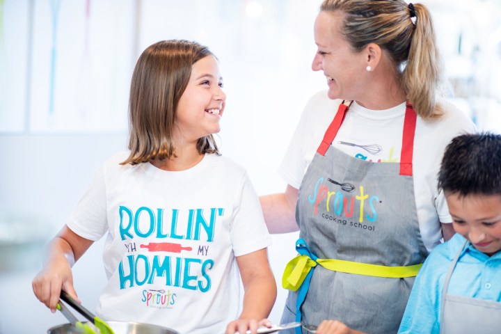 Woman and two children smiling in a kitchen setting, wearing aprons and engaging in a cooking activity.