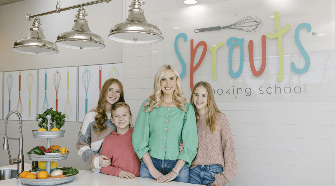 Four people smiling by a kitchen counter with fruit, under 'Sprouts Cooking School' sign.