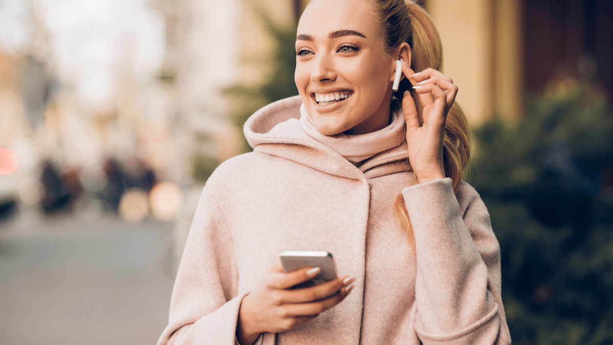 Woman in a pink coat smiling while using earbuds and holding a smartphone outdoors.