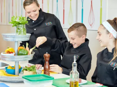 Three people in black chef coats cooking with vegetables, smiling in a kitchen.