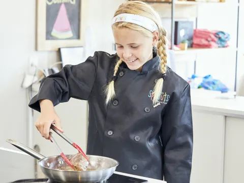Girl in chef coat cooking with tongs in a pan on a stovetop.