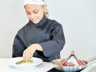 Child chef plating asparagus on rice, wearing black coat and headband in kitchen.