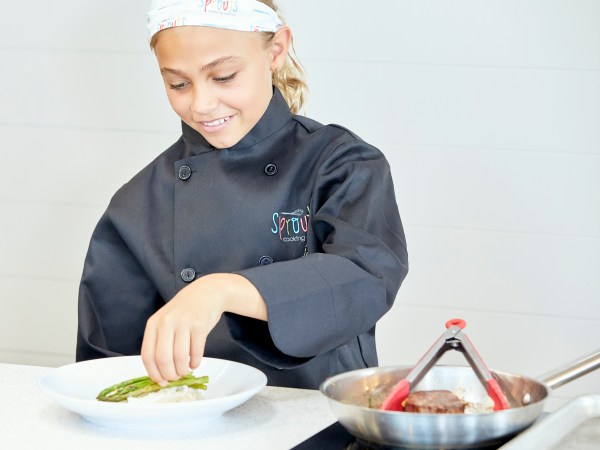 Child chef plating asparagus on rice, wearing black coat and headband in kitchen.