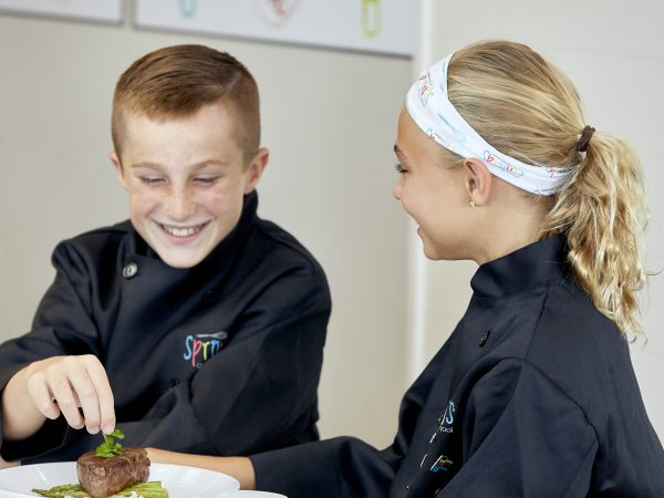 Two kids in chef coats, smiling while preparing a dish on a plate.
