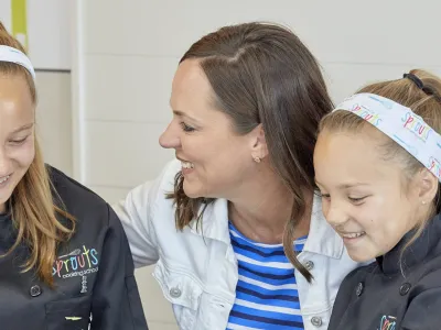 Two girls in chef coats and a woman smiling together at a table with a welcome sign and plated dish.