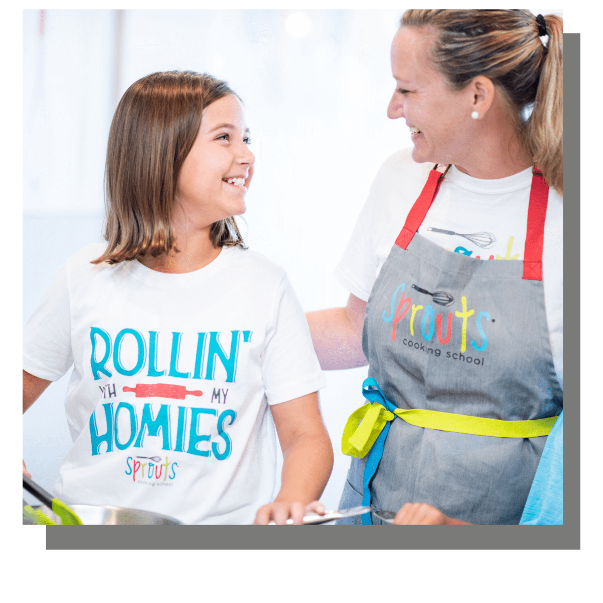Smiling woman and girl in Sprouts cooking school outfits looking at each other.