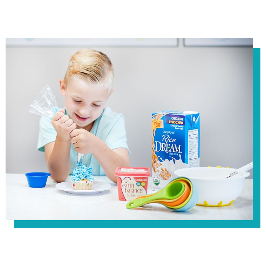Child decorating a cupcake with blue icing, surrounded by baking ingredients and utensils.