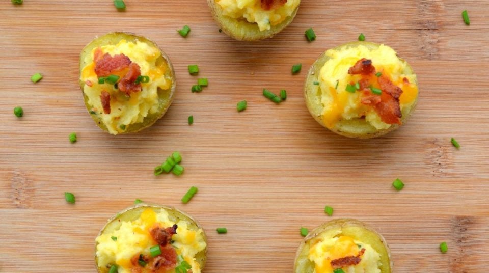 Five loaded potato halves with cheese, bacon, and chives on a wooden surface.