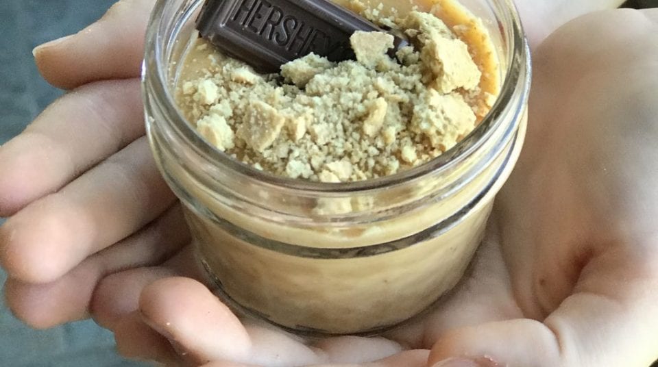 Hands holding a jar of dessert topped with crumbled cookies and a chocolate piece.