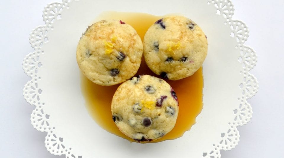 Three blueberry muffins on a white doily plate with maple syrup.