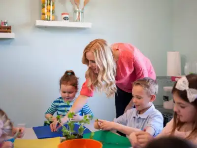 Woman assisting children with crafts at a table in a classroom.