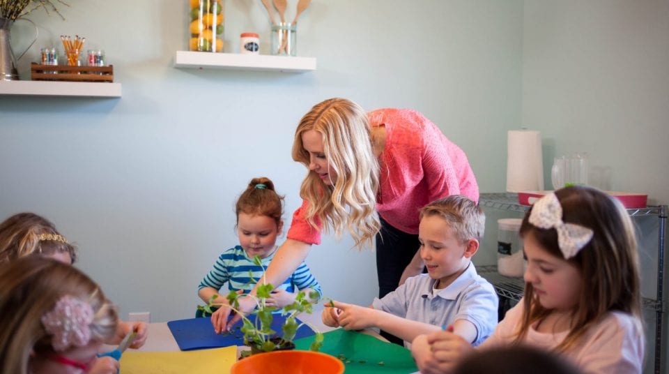 Woman assisting children with crafts at a table in a classroom.