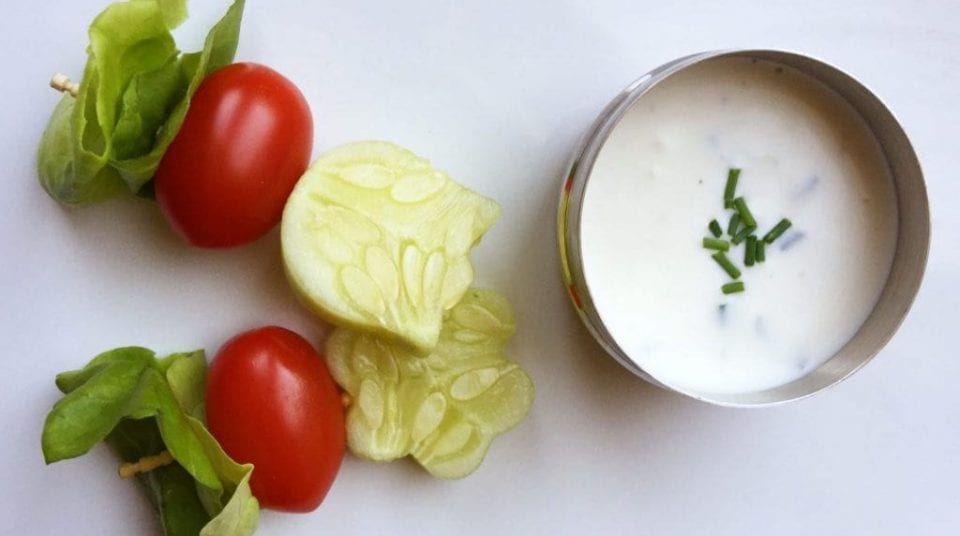 Cucumber and tomatoes on skewers beside a bowl of white sauce with chives.