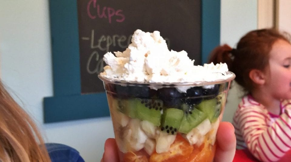Cup of fruit with whipped cream, hand holding it, child in background.
