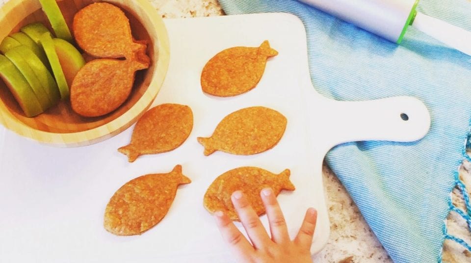 Child's hand reaching for fish-shaped cookies on a white board with apple slices in a bowl nearby.