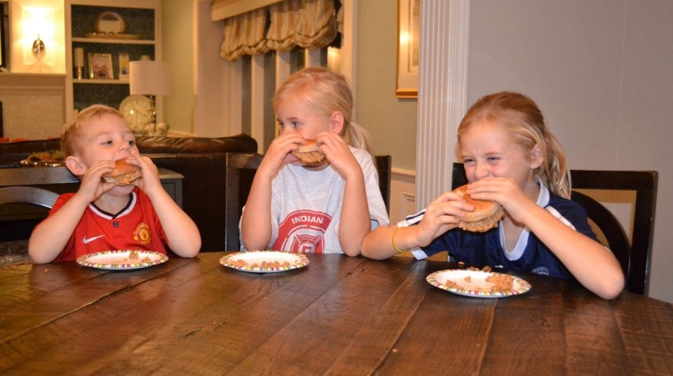 Three kids at a table eating burgers on paper plates in a cozy dining area.