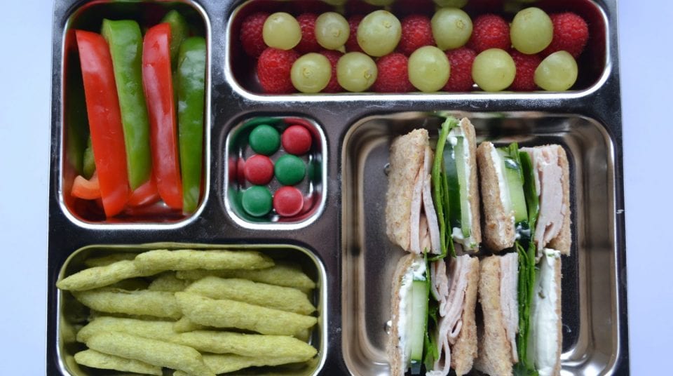 Stainless steel lunchbox with peppers, grapes, raspberries, candies, snacks, and sandwiches.