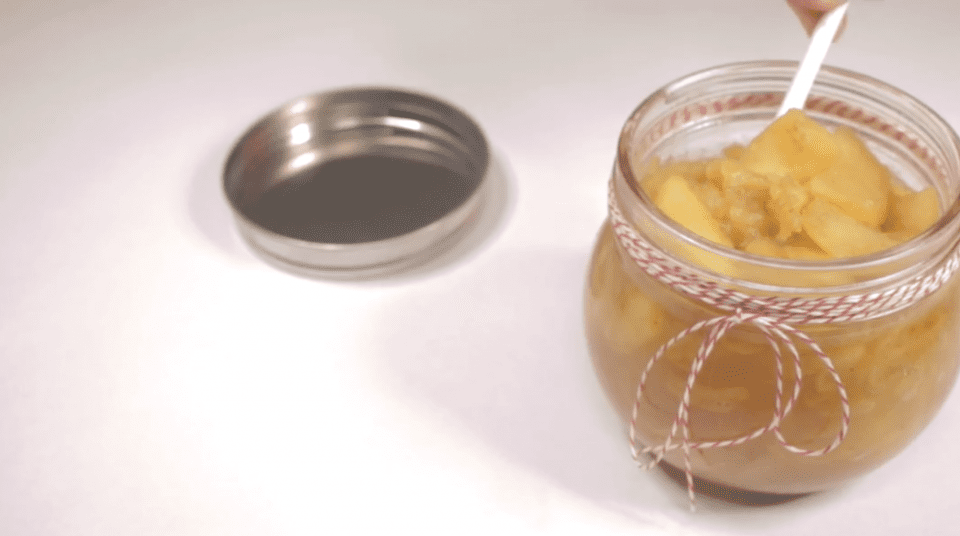 Open jar with yellow preserve and red-white string, lid next to it on white surface.