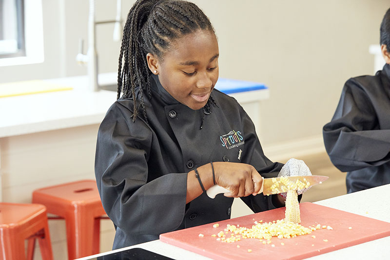 Young chef slicing corn in a kitchen, wearing a black apron.