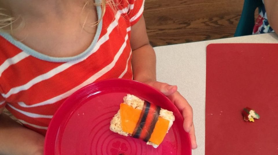 Child holding orange sushi-shaped treat on a red plate.