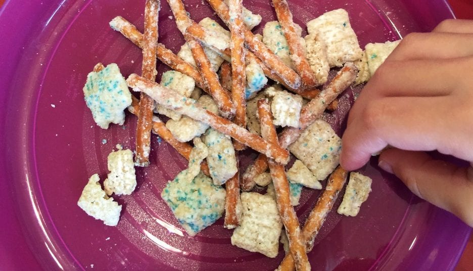 Snack mix of pretzels and cereal on a pink plate with a hand reaching in.