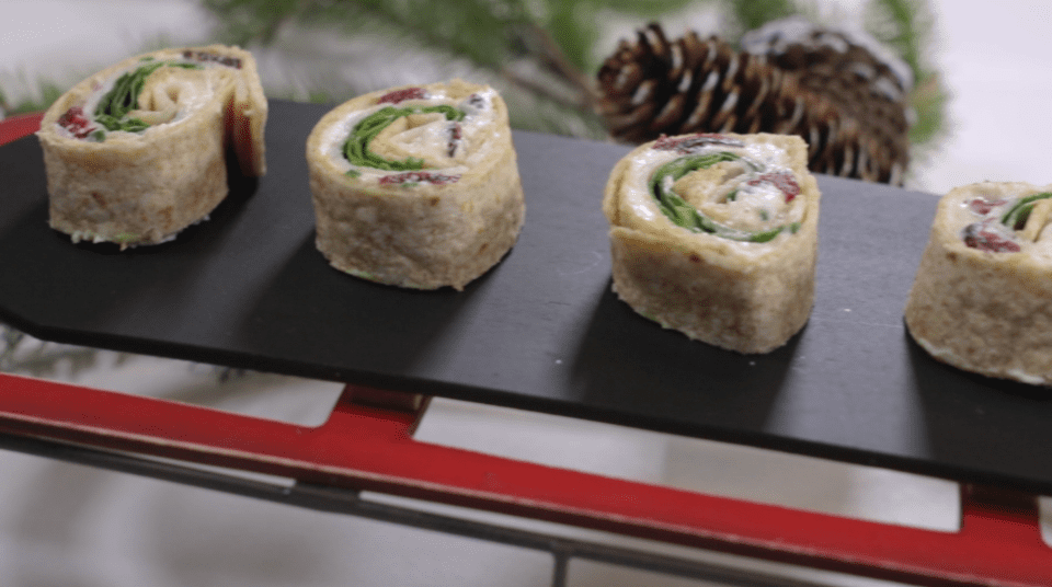 Four pinwheel sandwiches with greens on a black tray, pine cones in background.