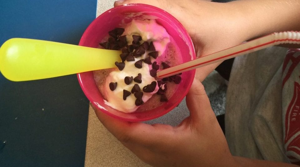 Close-up of hands holding a pink cup with ice cream, chocolate chips, a yellow spoon, and a straw.