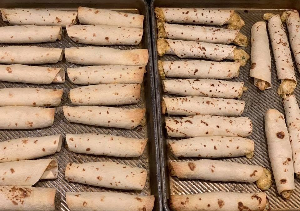 Two baking trays of rolled taquitos, some with visible filling, arranged neatly in rows.