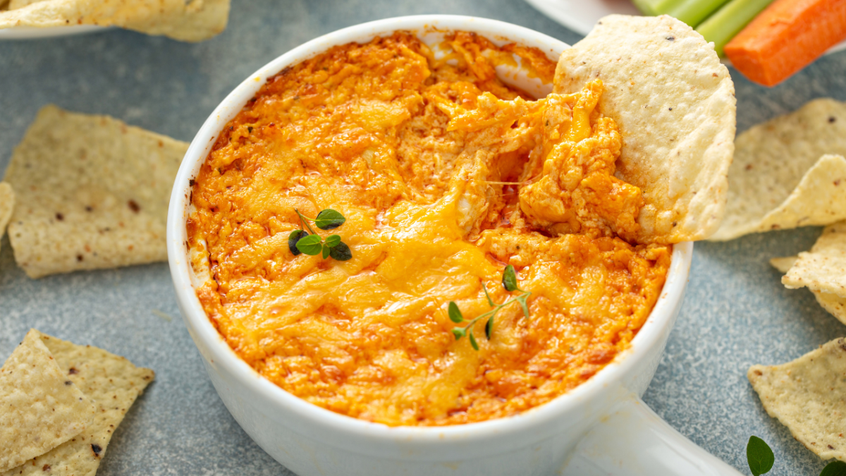 Cheesy buffalo chicken dip with chips in a white bowl, garnished with herbs.