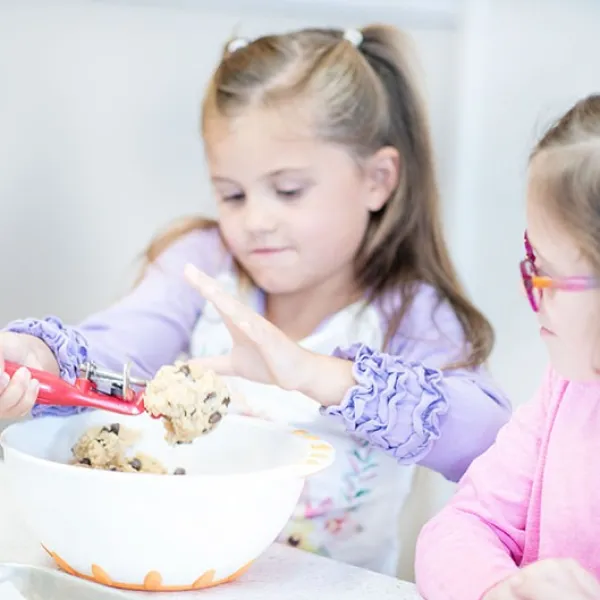 Two girls preparing cookie dough with a scoop in a mixing bowl.