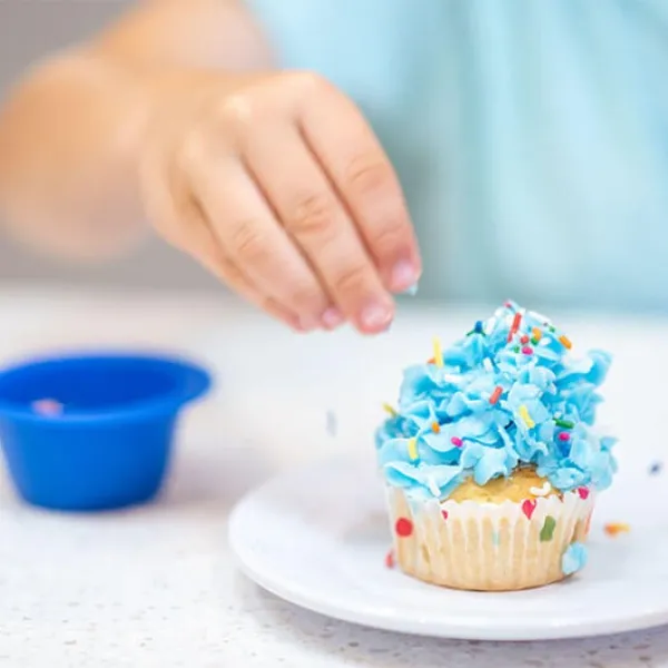 Child decorating cupcake with blue frosting and sprinkles on a plate.