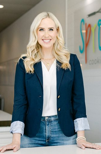 Smiling woman in blazer and jeans stands indoors, colorful logo on wall.