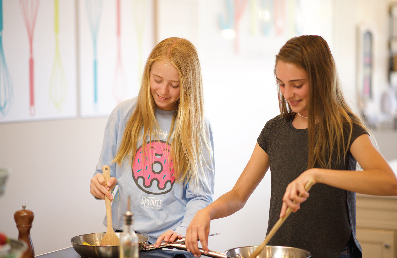 Two young girls cooking together, stirring pans with wooden spoons, in a bright kitchen.