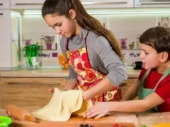 Two children in aprons roll out dough with a rolling pin in a kitchen.